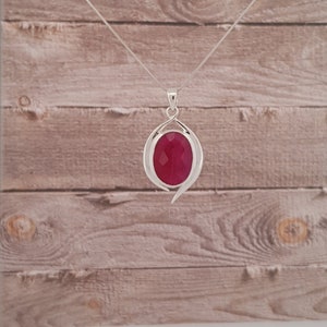 Op de afbeelding: Een zilveren ketting met een hanger met een grote, ovale, gefacetteerde, robijnrode edelsteen in een zilveren swirl-ontwerp.