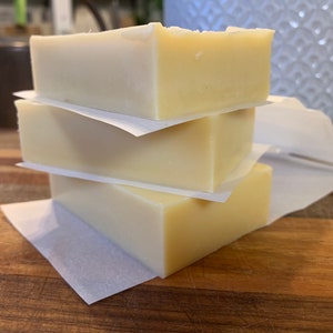 Pode incluir: Três barras de sabão branco embrulhadas em papel vegetal branco. O sabão está empilhado em uma superfície de madeira.
