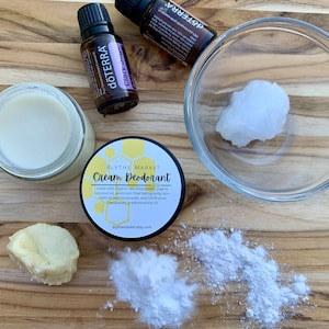 May include: A close-up of ingredients for making a homemade cream deodorant. The ingredients include shea butter, coconut oil, baking soda, arrowroot powder, and essential oil. The ingredients are on a wooden surface.