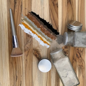 Puede incluir: Una superficie de madera con una variedad de polvos en pequeños frascos de vidrio. Los polvos están dispuestos en un patrón de arcoíris, con polvos negros, marrones, grises, beige, amarillos y blancos. También hay un pincel de maquillaje en la superficie.