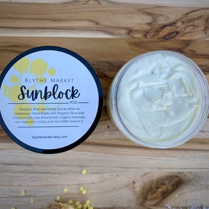 May include: A jar of white cream next to a container with a black lid. The lid has a label with the text "Sunblock" and "4OZ". The label also includes the text "BLYTHE MARKET". The cream is handmade with organic ingredients.