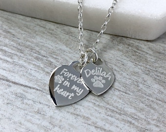 May include: A silver necklace with two heart-shaped pendants. One pendant reads "Forever in my heart" with paw prints, and the other says "Delilah" with a paw print. The chain is delicate and the pendants are shiny.