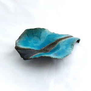 May include: A handmade ceramic dish with an organic shape. The dish has a vibrant turquoise interior, a dark grey stripe, and a textured, cracked surface. The edges are uneven, adding to its artistic appeal. The dish is likely 20cm in length.