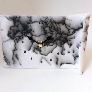 May include: A rectangular white clock with a black and gray abstract design. The clock face features black hands, a gold center, and navy blue hour markers. The clock has a unique, artistic appearance.