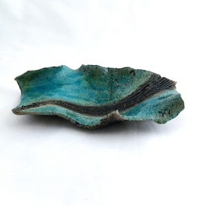 May include: A handmade ceramic dish with an irregular, natural form. The dish has a bright turquoise interior with a dark, textured band across the middle. The exterior has a mottled, earthy green-blue colour, creating a striking contrast. The dish is set against a white background.
