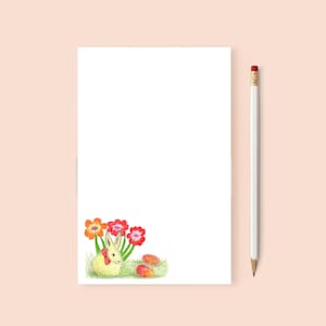 May include: A white bunny with a red bow sits in a field of green grass with orange and red flowers. There are three orange and red eggs scattered around the bunny.