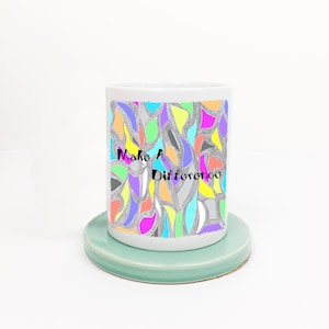 Peut inclure: Une tasse en céramique blanche avec un motif abstrait coloré et le texte "Make A Difference" imprimé sur le devant. La tasse est posée sur un dessous-de-tasse vert clair.