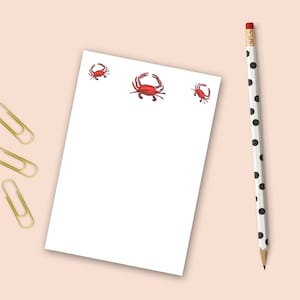 May include: A white notepad with three red crab illustrations. A black and white polka dot pencil is on the right side of the notepad. There are four gold paper clips on the left side of the notepad.