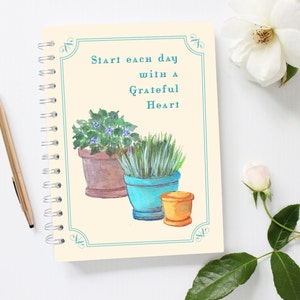 May include: A cream-colored spiral bound notebook with a teal border and the text "Start each day with a Grateful Heart". The notebook features a watercolor illustration of three potted plants: a green plant with blue flowers, a green grass plant, and a brown plant.