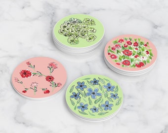 May include: Four round coasters with floral designs. The coasters are made of a white material with a glossy finish. The designs are in shades of pink, red, blue, and green.