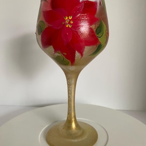 Pode incluir: Uma taça de vinho pintada à mão com um design vibrante de flor de poinsétia vermelha. A flor é acentuada com detalhes amarelos e folhas verdes. A haste e a base do copo têm um acabamento dourado, criando um visual elegante.