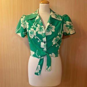 以下が含まれることがあります： 鮮やかな緑色に大きな白いハイビスカスの花柄がプリントされた、半袖のクロップド丈ボタンダウンシャツ。尖った襟と裾の前で結ぶリボンが特徴で、マネキンのトルソーに着用された状態で展示されています。