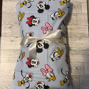 May include: A light blue fabric with a white ribbon tied around it. The fabric has a repeating pattern of Mickey Mouse, Minnie Mouse, Donald Duck, and Goofy.