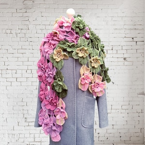 May include: A crocheted scarf with pink, green, and yellow flowers. The scarf is draped over a gray coat on a white brick wall background.