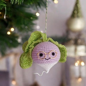 May include: A crocheted purple turnip with a white bottom and green leaves, wearing gold-rimmed glasses and a smiling face. The turnip is hanging from a string on a Christmas tree.