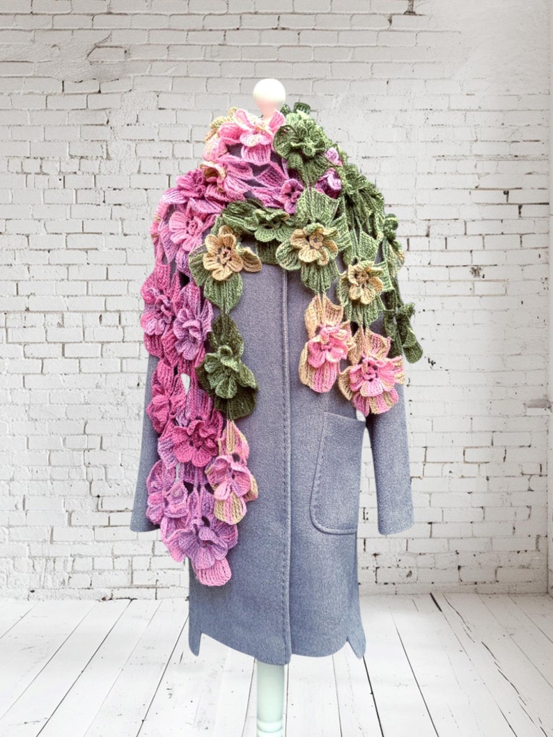 May include: A crocheted scarf with pink, green, and yellow flowers. The scarf is draped over a gray coat on a white brick wall background.