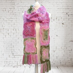 May include: A handmade crochet scarf in shades of pink and green, displayed on a mannequin. The scarf features intricate floral patterns and a fringed edge. The scarf is approximately 72 inches long.