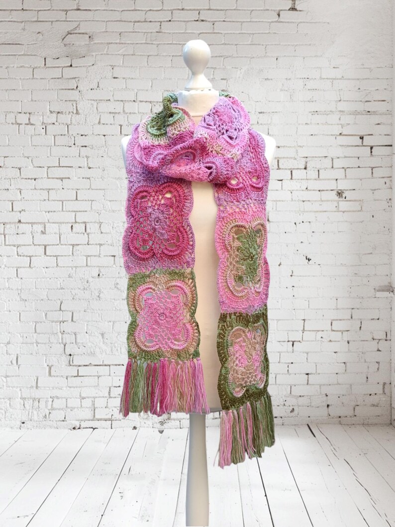 May include: A handmade crochet scarf in shades of pink and green, displayed on a mannequin. The scarf features intricate floral patterns and a fringed edge. The scarf is approximately 72 inches long.