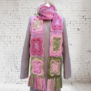 May include: A handmade crochet scarf in shades of pink, green, and cream, displayed on a mannequin. The scarf features a repeating square pattern with fringe at the ends. The scarf is draped over a gray coat.