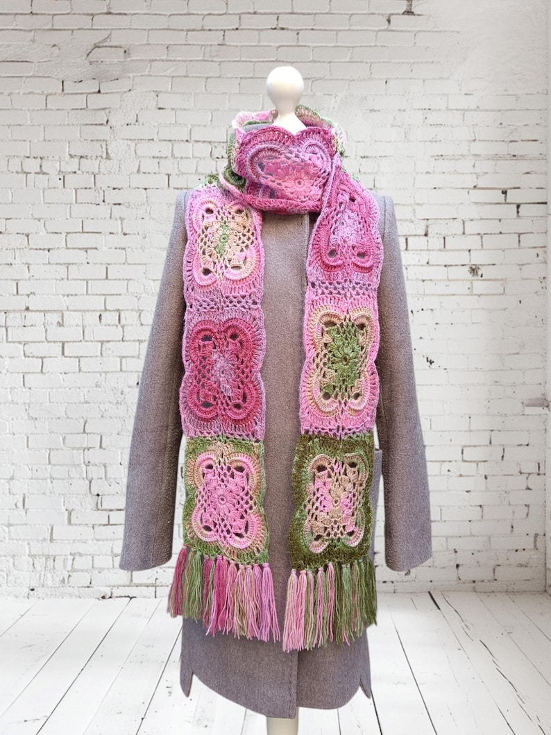 May include: A handmade crochet scarf in shades of pink, green, and cream, displayed on a mannequin. The scarf features a repeating square pattern with fringe at the ends. The scarf is draped over a gray coat.