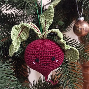 May include: A crocheted beetroot ornament with a smiling face, green leaves, and a red body. The ornament is hanging on a green Christmas tree.