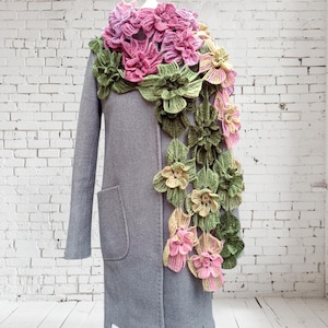 May include: A crocheted scarf with pink, green, and yellow flowers. The scarf is draped over a gray coat on a white brick wall background.