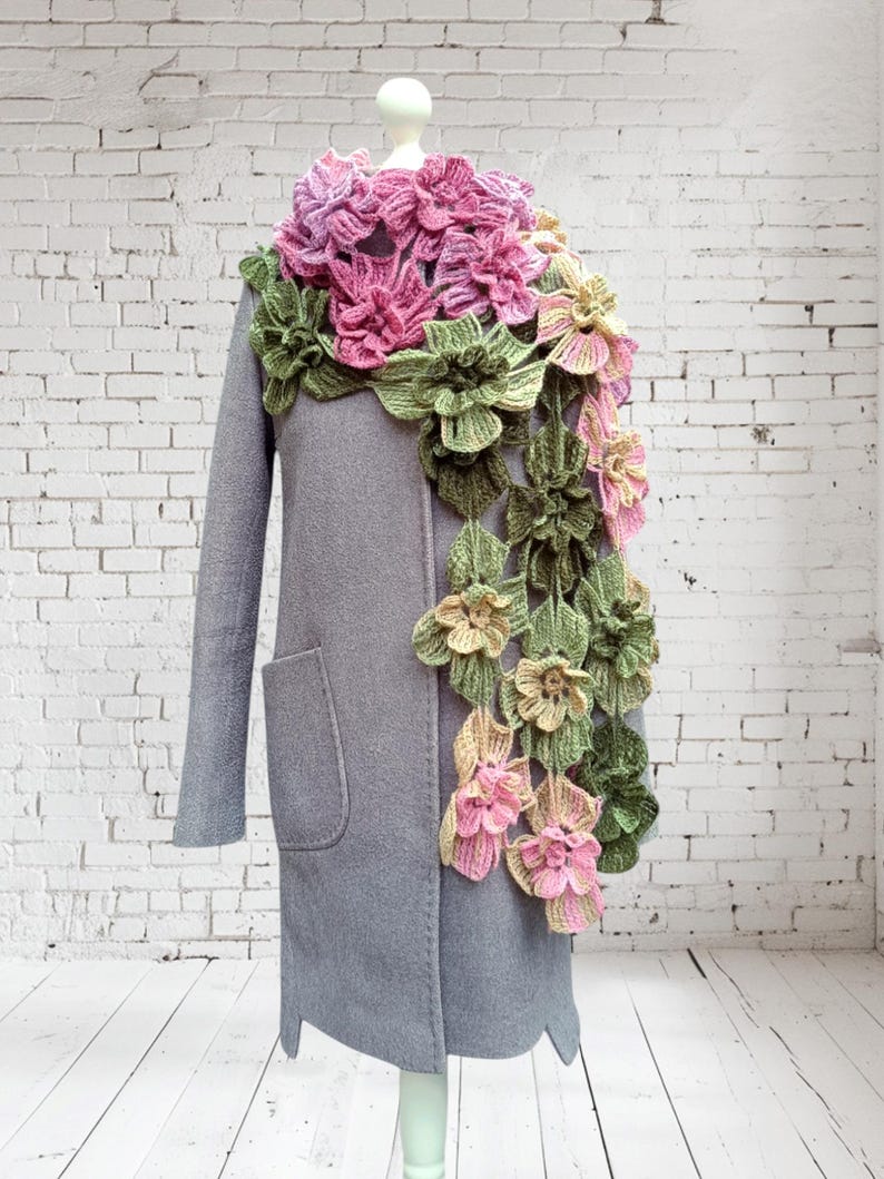 May include: A crocheted scarf with pink, green, and yellow flowers. The scarf is draped over a gray coat on a white brick wall background.