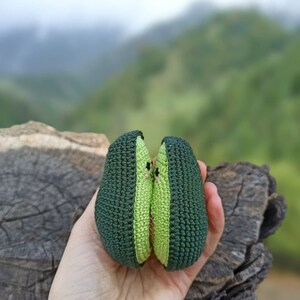 May include: Two crocheted avocado halves, green with a light green center, are held together in a hand. The avocados have a cute face with two small pink dots for eyes.