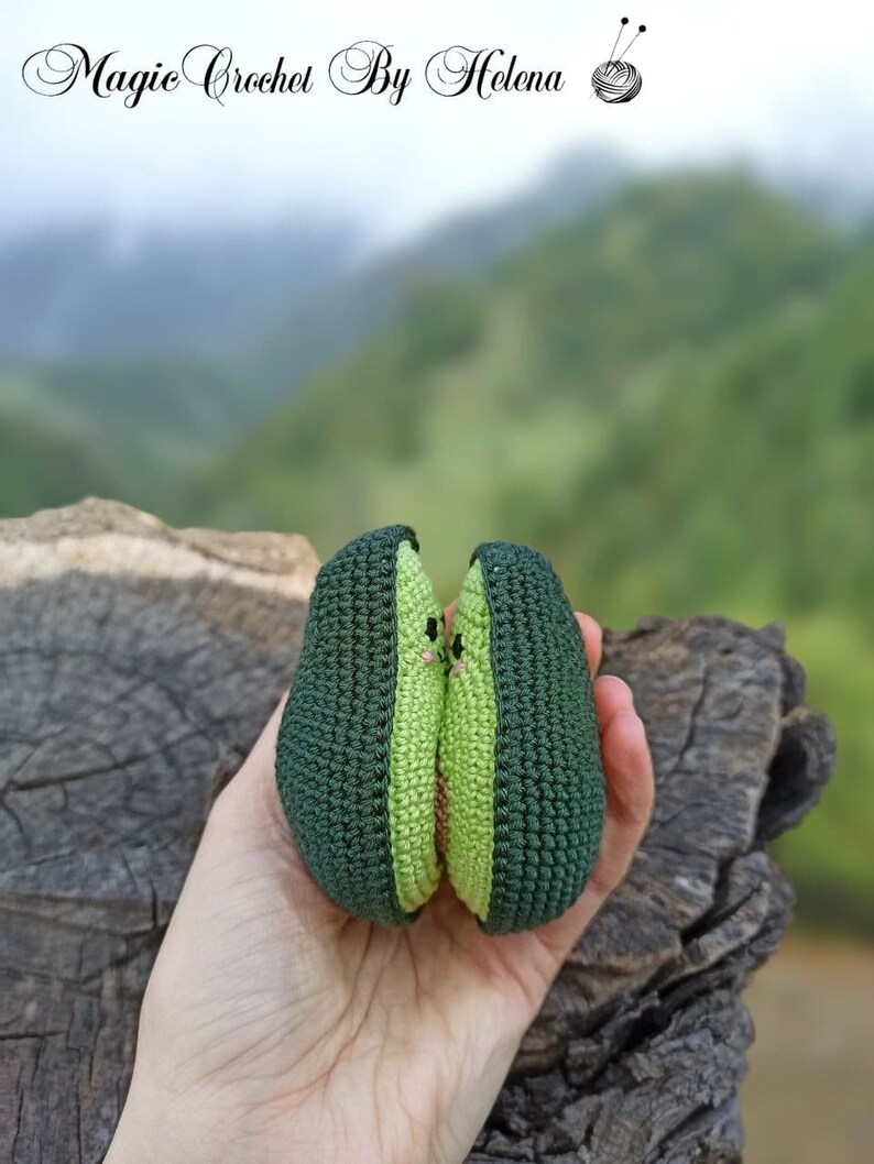 May include: Two crocheted avocado halves, green with a light green center, are held together in a hand. The avocados have a cute face with two small pink dots for eyes.