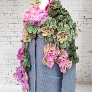 May include: A crocheted scarf with pink, green, and beige flowers. The scarf is draped over a gray coat.
