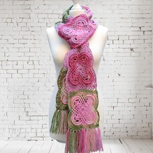 May include: A handmade crochet scarf in shades of pink and green, displayed on a mannequin. The scarf features intricate floral patterns and a fringed edge. The color palette includes various shades of pink, with accents of green.