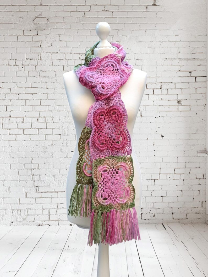 May include: A handmade crochet scarf in shades of pink and green, displayed on a mannequin. The scarf features intricate floral patterns and a fringed edge. The color palette includes various shades of pink, with accents of green.
