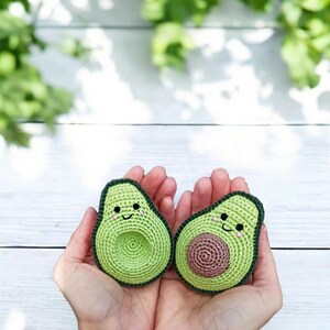 May include: Two crocheted avocado halves with smiling faces. The avocado halves are green with brown pits and are held in a person's hands.