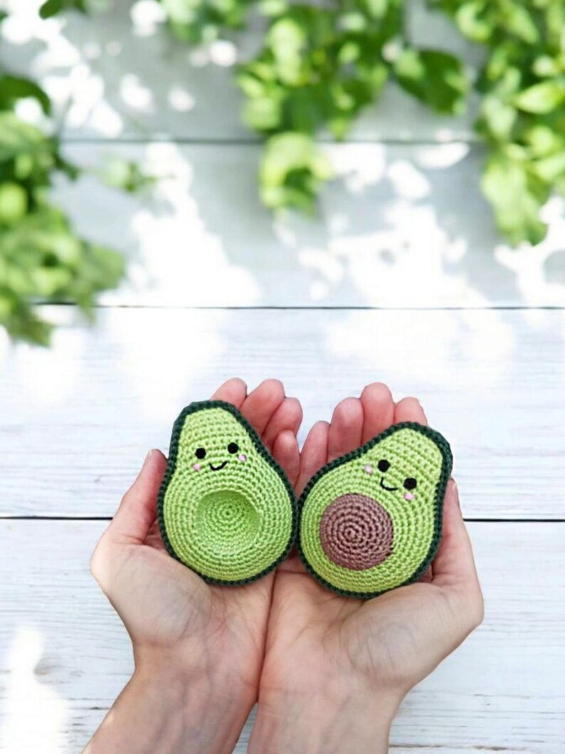 May include: Two crocheted avocado halves with smiling faces. The avocado halves are green with brown pits and are held in a person's hands.