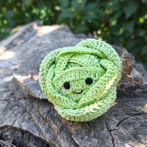 May include: A green crocheted cabbage with a smiling face. The cabbage is sitting on a wooden surface.