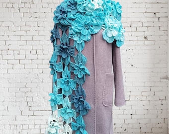 Puede incluir: Un abrigo lavanda se exhibe en un maniquí blanco, adornado con una bufanda de ganchillo. La bufanda presenta una cascada de flores azules y blancas, creando un contraste llamativo con el color neutro del abrigo. El diseño floral añade un toque de encanto artesanal.
