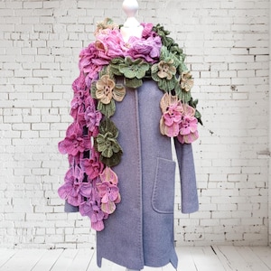 May include: A crocheted scarf with pink, green, and beige flowers. The scarf is draped over a mannequin wearing a gray coat.