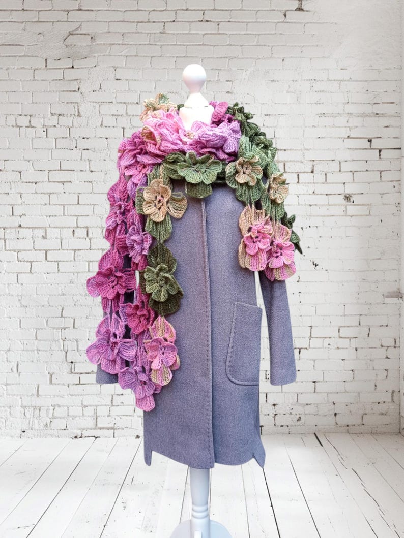 May include: A crocheted scarf with pink, green, and beige flowers. The scarf is draped over a mannequin wearing a gray coat.