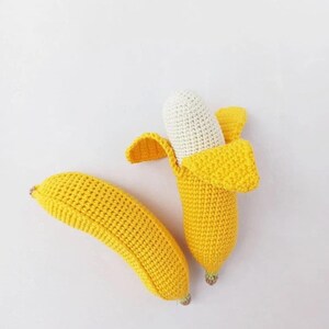 Puede incluir: Dos juguetes de pl&aacute;tano tejidos a crochet. Un pl&aacute;tano est&aacute; entero y el otro est&aacute; partido para revelar un interior tejido a crochet blanco.