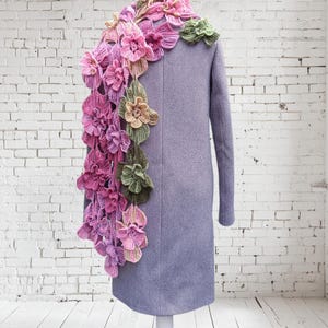 May include: A purple coat with a crocheted flower scarf in shades of pink, green, and yellow. The scarf is draped over the shoulder and down the back of the coat.