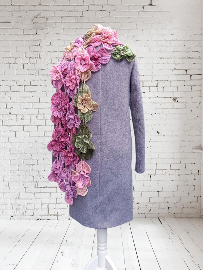 May include: A purple coat with a crocheted flower scarf in shades of pink, green, and yellow. The scarf is draped over the shoulder and down the back of the coat.