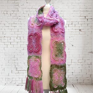 May include: A handmade crochet scarf in shades of pink, green, and olive. The scarf features a floral design and fringe tassels at the ends. The scarf is displayed on a white mannequin against a white brick wall.