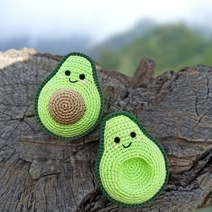May include: Two crocheted avocado plushies with smiling faces. The avocados are green with brown pits and are sitting on a wooden surface.