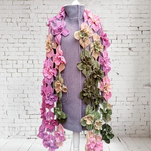 May include: A crocheted scarf with pink, green, and yellow flowers. The scarf is draped over a white mannequin.