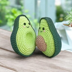 May include: Two crocheted avocado plush toys with smiling faces. The avocados are green with brown pits and are sitting on a wooden surface.