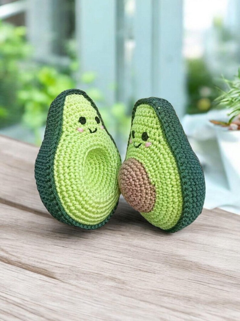 May include: Two crocheted avocado plush toys with smiling faces. The avocados are green with brown pits and are sitting on a wooden surface.