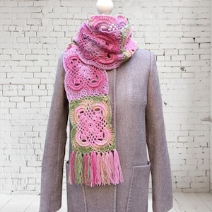 May include: A pink, green, and white crocheted scarf with a floral design and fringe. The scarf is draped over a gray coat on a mannequin. The scarf's colors include shades of pink, green, and white.