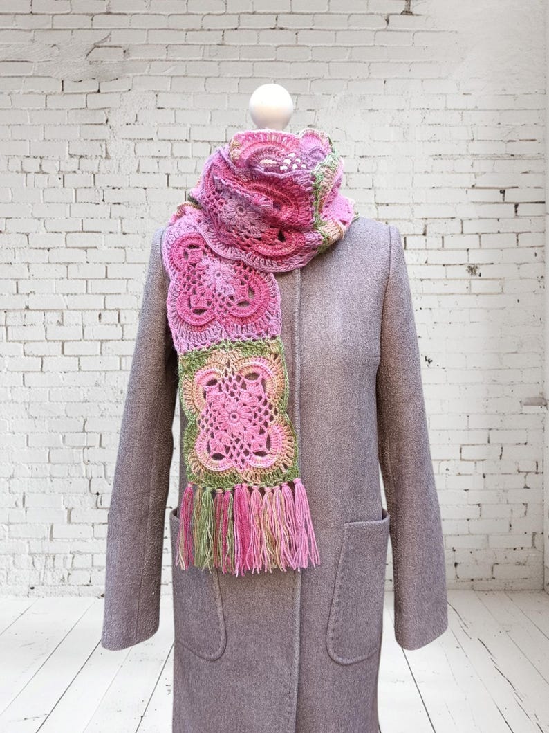 May include: A pink, green, and white crocheted scarf with a floral design and fringe. The scarf is draped over a gray coat on a mannequin. The scarf's colors include shades of pink, green, and white.