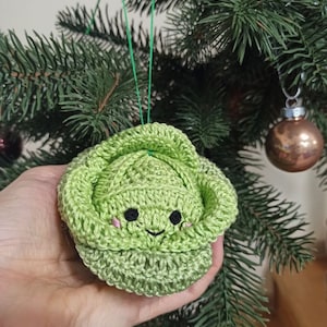 Könnte beinhalten: Ein grüner, gehäkelter Weihnachtsbaumschmuck in Form eines Kohlkopfes mit einem lächelnden Gesicht. Der Schmuck hängt an einem grünen Weihnachtsbaum.