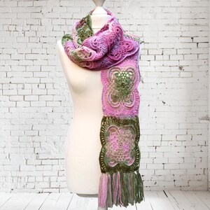 May include: A handmade crochet scarf in shades of pink, green, and olive. The scarf features a detailed floral pattern and a fringed edge. The scarf is displayed on a white mannequin against a white brick wall.